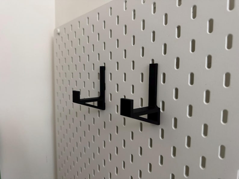 Op de afbeelding: Twee zwarte metalen pegboardhaken zijn gemonteerd op een witte pegboard. De haken zijn L-vormig en ontworpen om items vast te houden. Het pegboard heeft een raster van gelijkmatig verdeelde gaten.