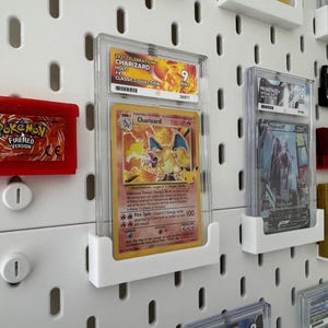 Może przedstawiać: Biała tablica z gwoździami z r&oacute;żnymi kartami kolekcjonerskimi i kasetami do gier Pokemon. Karty są w ochronnych plastikowych etui i są zamontowane na białych plastikowych uchwytach. Kasety są w czerwonych i złotych obudowach. Tablica z gwoździami ma siatkę otwor&oacute;w.