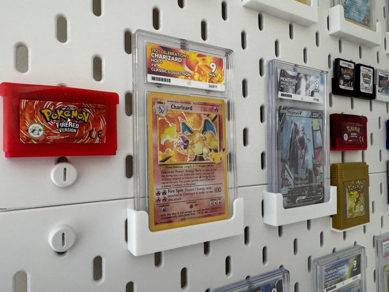 Może przedstawiać: Biała tablica z gwoździami z r&oacute;żnymi kartami kolekcjonerskimi i kasetami do gier Pokemon. Karty są w ochronnych plastikowych etui i są zamontowane na białych plastikowych uchwytach. Kasety są w czerwonych i złotych obudowach. Tablica z gwoździami ma siatkę otwor&oacute;w.
