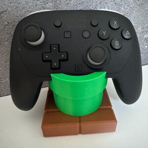 K&ouml;nnte beinhalten: Schwarzer Videospiel-Controller auf einem gr&uuml;nen Rohr- und braunen Ziegelst&auml;nder. Der Controller hat wei&szlig;e Akzente und Tasten mit den Bezeichnungen X, Y, A und B. Der St&auml;nder ist ein dekorativer Artikel.