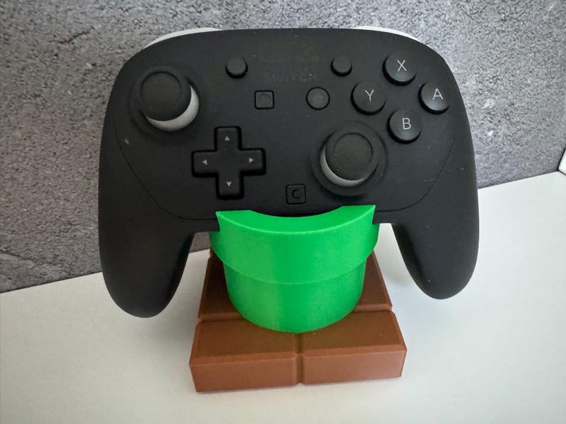 K&ouml;nnte beinhalten: Schwarzer Videospiel-Controller auf einem gr&uuml;nen Rohr- und braunen Ziegelst&auml;nder. Der Controller hat wei&szlig;e Akzente und Tasten mit den Bezeichnungen X, Y, A und B. Der St&auml;nder ist ein dekorativer Artikel.