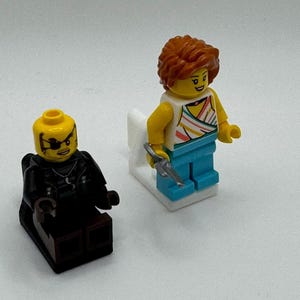 K&ouml;nnte beinhalten: Zwei Lego-Minifiguren. Eine Figur sitzt und tr&auml;gt ein schwarzes Outfit und eine Augenklappe. Die andere Figur steht und tr&auml;gt ein wei&szlig;es Oberteil mit bunten Streifen, eine blaue Hose und h&auml;lt einen silbernen Gegenstand.