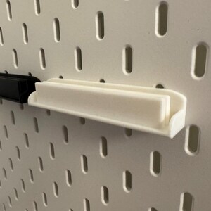 Pode incluir: Duas pequenas prateleiras retangulares, uma preta e outra branca, est&atilde;o montadas numa placa perfurada branca. As prateleiras t&ecirc;m um design simples e minimalista, com uma base plana e uma borda elevada para segurar objetos.