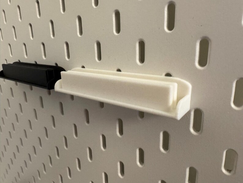 Pode incluir: Duas pequenas prateleiras retangulares, uma preta e outra branca, est&atilde;o montadas numa placa perfurada branca. As prateleiras t&ecirc;m um design simples e minimalista, com uma base plana e uma borda elevada para segurar objetos.