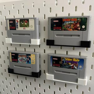 May include: Four gray Super Nintendo game cartridges, each with a different game label, mounted on a white pegboard. Games include soccer, Donkey Kong Country, Super Mario All-Stars, and Stunt Race. The cartridges are held in place by white and black holders.
