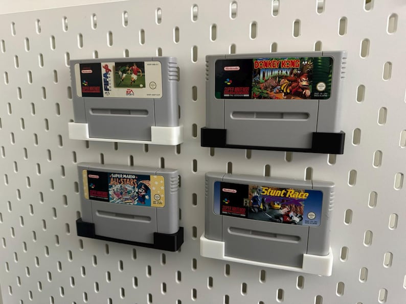 May include: Four gray Super Nintendo game cartridges, each with a different game label, mounted on a white pegboard. Games include soccer, Donkey Kong Country, Super Mario All-Stars, and Stunt Race. The cartridges are held in place by white and black holders.