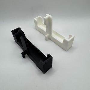May include: Two 3D-printed objects. One is black, the other is white. Both have a rectangular base with a raised section and a vertical support. The objects appear to be designed to hold something.
