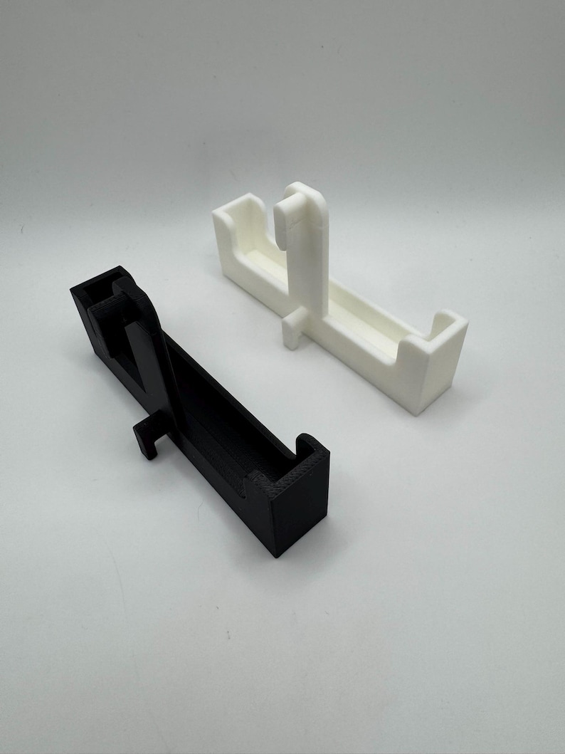 May include: Two 3D-printed objects. One is black, the other is white. Both have a rectangular base with a raised section and a vertical support. The objects appear to be designed to hold something.