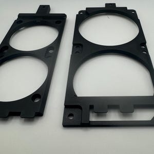 K&ouml;nnte beinhalten: Zwei schwarze Kunststoff-L&uuml;fterhalterungen mit runden Ausschnitten f&uuml;r L&uuml;fter. Die Halterungen haben eine strukturierte Oberfl&auml;che und sind f&uuml;r die Befestigung an einer Oberfl&auml;che konzipiert.