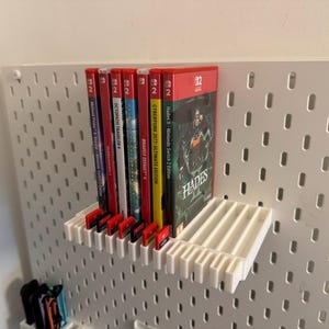 May include: A white shelf holds several Nintendo Switch game cases, including "Hades II," "Cyberpunk 2077," and "Octopath Traveler." The games are displayed against a white pegboard wall, with game cartridges visible below.
