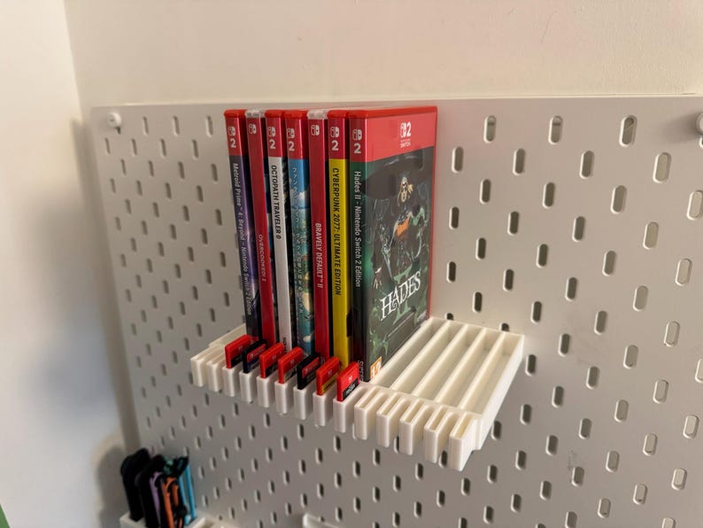 May include: A white shelf holds several Nintendo Switch game cases, including "Hades II," "Cyberpunk 2077," and "Octopath Traveler." The games are displayed against a white pegboard wall, with game cartridges visible below.