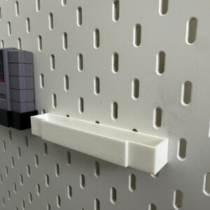 K&ouml;nnte beinhalten: Eine graue Super Nintendo-Spielkassette wird auf einem schwarzen Regal gezeigt, das an einer wei&szlig;en Lochwand befestigt ist. Ein wei&szlig;es Regal ist ebenfalls an der Lochwand montiert.