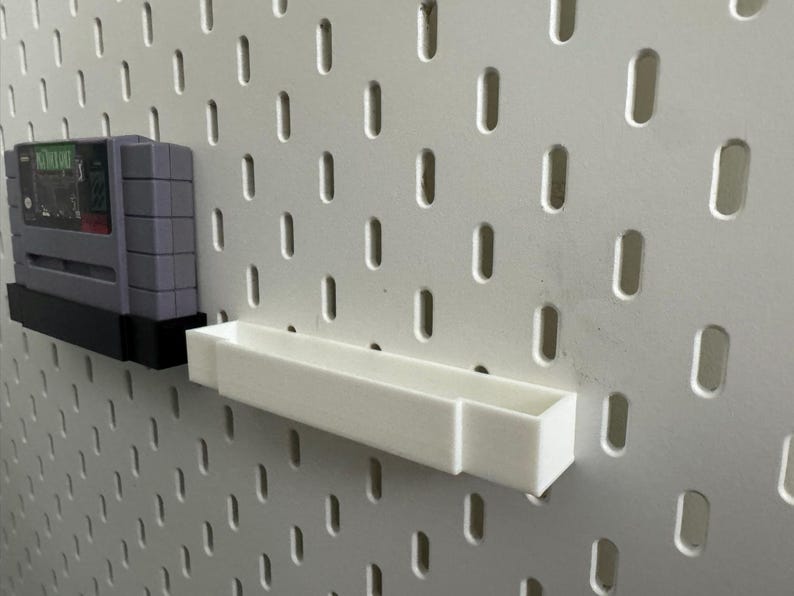 K&ouml;nnte beinhalten: Eine graue Super Nintendo-Spielkassette wird auf einem schwarzen Regal gezeigt, das an einer wei&szlig;en Lochwand befestigt ist. Ein wei&szlig;es Regal ist ebenfalls an der Lochwand montiert.