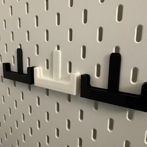 Op de afbeelding: Drie pegboard haken in zwart en wit. De haken hebben de vorm van een "T" met een platte basis en een verticale steun. Ze zijn ontworpen om in de gaten van een pegboard te passen. Het pegboard is wit.
