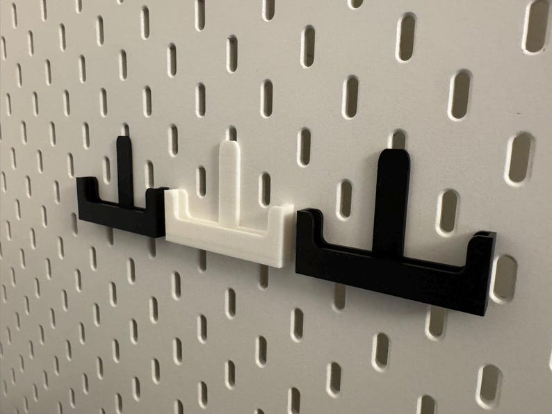 Op de afbeelding: Drie pegboard haken in zwart en wit. De haken hebben de vorm van een "T" met een platte basis en een verticale steun. Ze zijn ontworpen om in de gaten van een pegboard te passen. Het pegboard is wit.