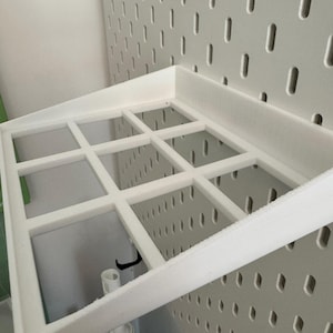 Op de afbeelding: Wit plasticen plank met een rasterpatroon, gemonteerd op een witte prikbordwand.