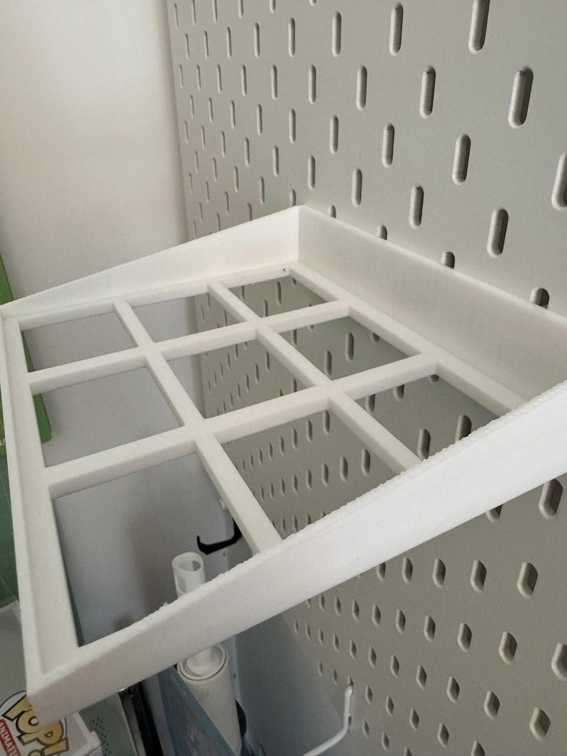 Op de afbeelding: Wit plasticen plank met een rasterpatroon, gemonteerd op een witte prikbordwand.