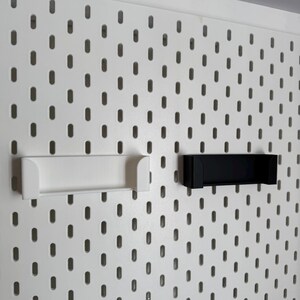K&ouml;nnte beinhalten: Wei&szlig;e und schwarze Zubeh&ouml;rteile f&uuml;r Lochw&auml;nde, darunter ein wei&szlig;es Regal und ein schwarzes Regal, sind an einer wei&szlig;en Lochwand befestigt. Die Lochwand hat gleichm&auml;&szlig;ig verteilte ovale L&ouml;cher. Die Zubeh&ouml;rteile sind zum Halten von Gegenst&auml;nden konzipiert.