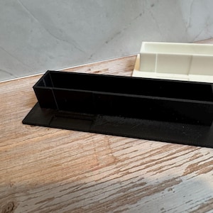 K&ouml;nnte beinhalten: Ein schwarzer rechteckiger Beh&auml;lter mit erh&ouml;htem Sockel, wahrscheinlich 3D-gedruckt, steht auf einer Holzoberfl&auml;che. Ein zweiter, wei&szlig;er rechteckiger Beh&auml;lter ist im Hintergrund zu sehen. Die Beh&auml;lter dienen wahrscheinlich zur Organisation kleiner Gegenst&auml;nde.