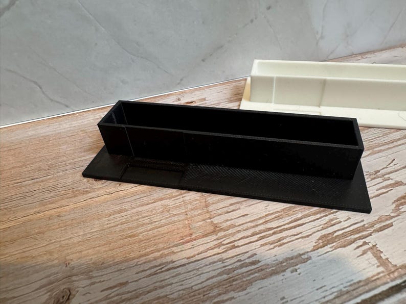 K&ouml;nnte beinhalten: Ein schwarzer rechteckiger Beh&auml;lter mit erh&ouml;htem Sockel, wahrscheinlich 3D-gedruckt, steht auf einer Holzoberfl&auml;che. Ein zweiter, wei&szlig;er rechteckiger Beh&auml;lter ist im Hintergrund zu sehen. Die Beh&auml;lter dienen wahrscheinlich zur Organisation kleiner Gegenst&auml;nde.