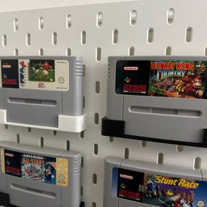May include: Four grey Super Nintendo game cartridges are displayed on a white pegboard. The cartridges are labelled with game titles such as "EA Sports," "Donkey Kong Country," "Super Mario All-Stars," and "Stunt Race FX."