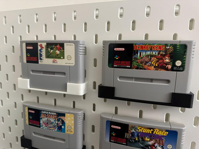 May include: Four grey Super Nintendo game cartridges are displayed on a white pegboard. The cartridges are labelled with game titles such as "EA Sports," "Donkey Kong Country," "Super Mario All-Stars," and "Stunt Race FX."