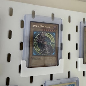 Pu&ograve; includere: Un pannello forato bianco con un portacarte in plastica trasparente che mostra una carta da gioco "Dark Magician". La carta presenta un personaggio di mago oscuro con uno staff e un cerchio di magia oscura. La carta &egrave; in una custodia protettiva.
