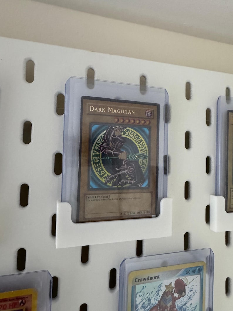 Pu&ograve; includere: Un pannello forato bianco con un portacarte in plastica trasparente che mostra una carta da gioco "Dark Magician". La carta presenta un personaggio di mago oscuro con uno staff e un cerchio di magia oscura. La carta &egrave; in una custodia protettiva.