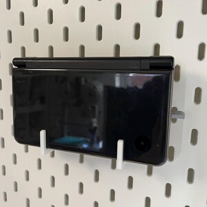 K&ouml;nnte beinhalten: Eine schwarze Handheld-Spielkonsole ist mit wei&szlig;en Halterungen an einer wei&szlig;en Lochwand befestigt. Die Konsole befindet sich in geschlossenem Zustand und zeigt ihre glatte, reflektierende Oberfl&auml;che. Die Lochwand hat gleichm&auml;&szlig;ig verteilte ovale L&ouml;cher.