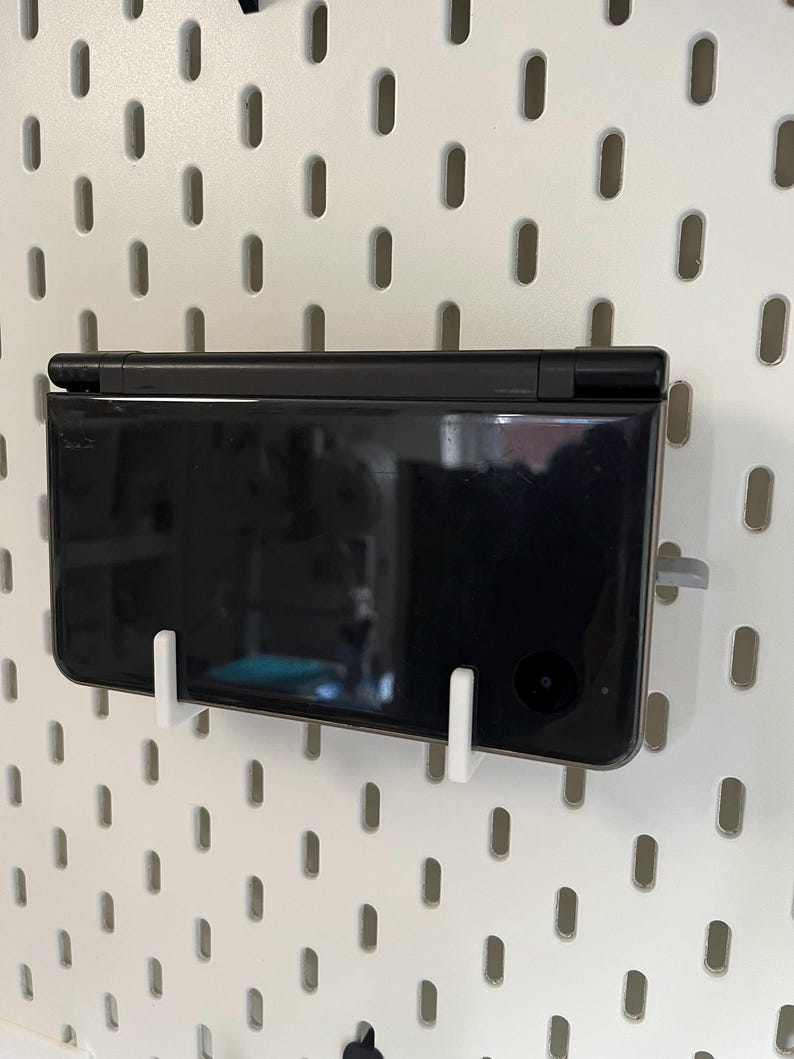 K&ouml;nnte beinhalten: Eine schwarze Handheld-Spielkonsole ist mit wei&szlig;en Halterungen an einer wei&szlig;en Lochwand befestigt. Die Konsole befindet sich in geschlossenem Zustand und zeigt ihre glatte, reflektierende Oberfl&auml;che. Die Lochwand hat gleichm&auml;&szlig;ig verteilte ovale L&ouml;cher.