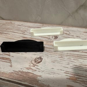 May include: Three small, rectangular, 3D-printed objects. One is black, and two are white. The objects have a curved top and a recessed area. They are arranged on a light-colored, wooden surface.
