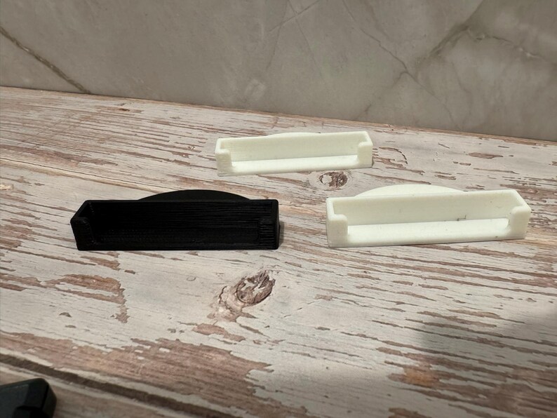 May include: Three small, rectangular, 3D-printed objects. One is black, and two are white. The objects have a curved top and a recessed area. They are arranged on a light-colored, wooden surface.