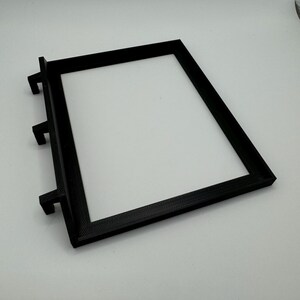 May include: Black plastic frame with three tabs on the side. The frame is designed to hold a small object, such as a picture or a piece of paper.