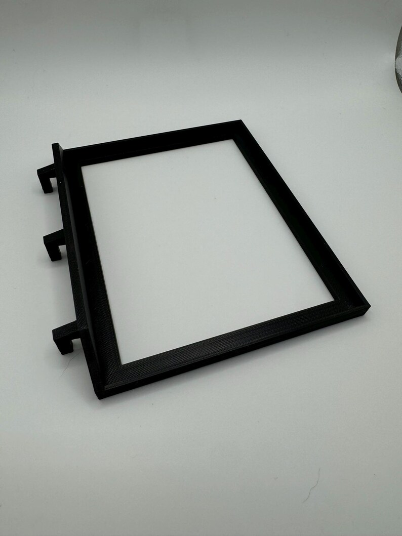 May include: Black plastic frame with three tabs on the side. The frame is designed to hold a small object, such as a picture or a piece of paper.