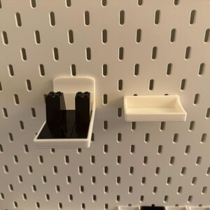 May include: White plastic pegboard accessories, including a shelf and a holder with black inserts. The items are designed to attach to a white pegboard with evenly spaced holes. The shelf is rectangular and the holder has a unique shape.