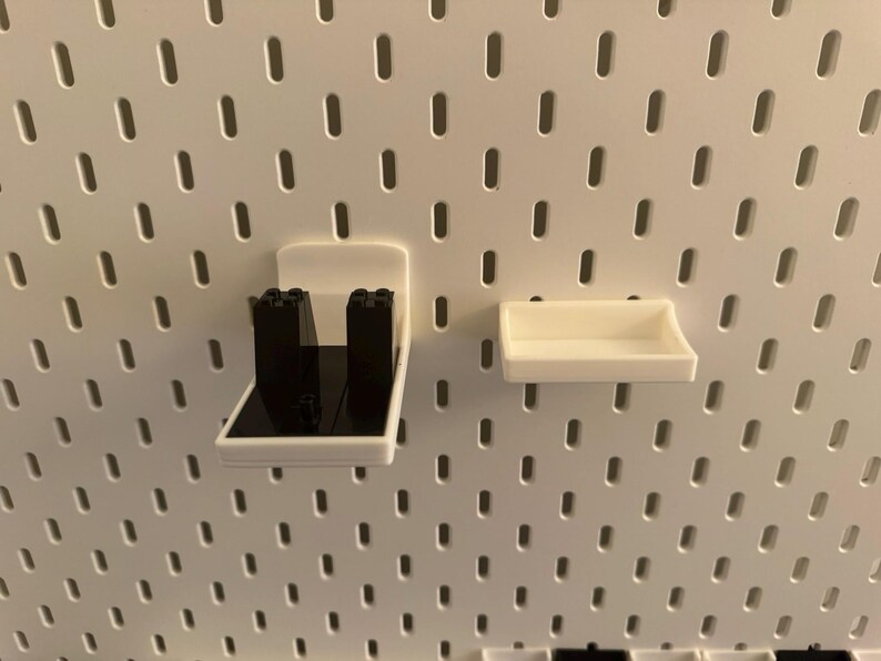 May include: White plastic pegboard accessories, including a shelf and a holder with black inserts. The items are designed to attach to a white pegboard with evenly spaced holes. The shelf is rectangular and the holder has a unique shape.
