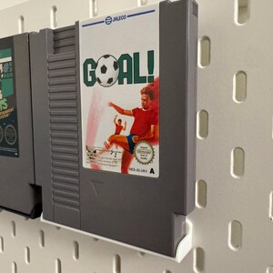 Pode incluir: Dois cartuchos de videojogos retro, um com a etiqueta "Tennis Entertainment System" e o outro "GOAL!" com tema de futebol. Os cartuchos s&atilde;o cinzentos e est&atilde;o montados numa parede perfurada branca.