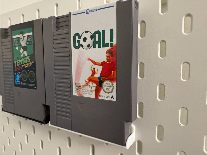 Pode incluir: Dois cartuchos de videojogos retro, um com a etiqueta "Tennis Entertainment System" e o outro "GOAL!" com tema de futebol. Os cartuchos s&atilde;o cinzentos e est&atilde;o montados numa parede perfurada branca.