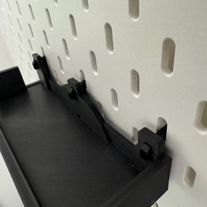 May include: Black wall-mounted shelf with a metal bracket and a pegboard background.