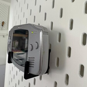 May include: A silver Game Boy Advance handheld console mounted on a white pegboard with a black bracket. The screen is visible, and the console's buttons are on the right side. The text "GAME BOY ADVANCE" is printed on the bottom.