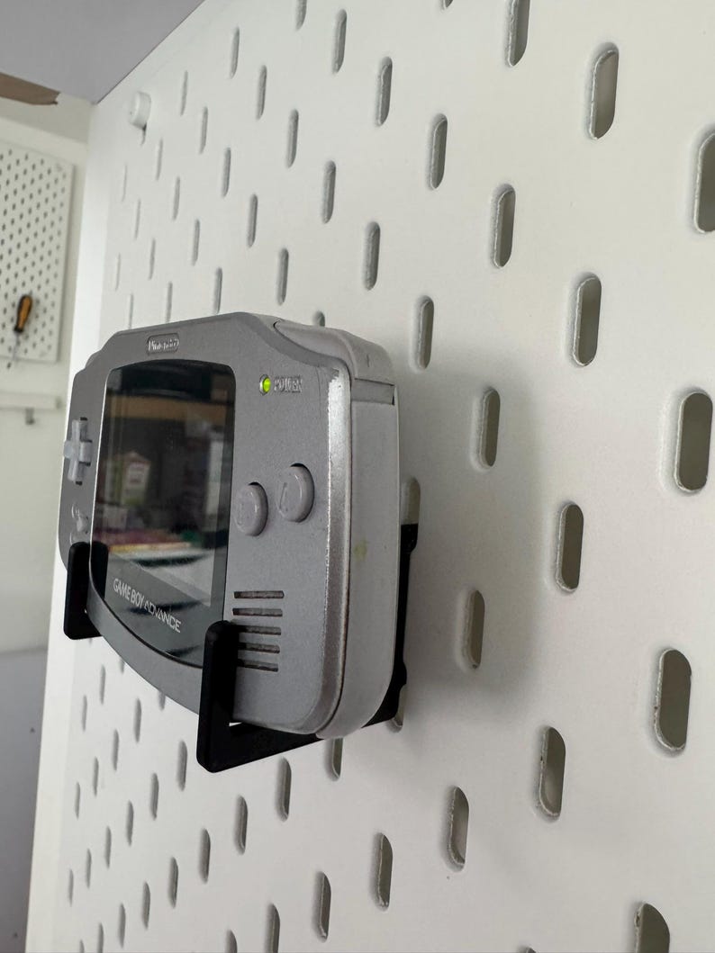 May include: A silver Game Boy Advance handheld console mounted on a white pegboard with a black bracket. The screen is visible, and the console's buttons are on the right side. The text "GAME BOY ADVANCE" is printed on the bottom.