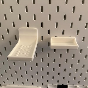 May include: Two white plastic pegboard accessories: a curved shelf with a dotted pattern and a small rectangular shelf. The items are mounted on a white pegboard with evenly spaced holes.