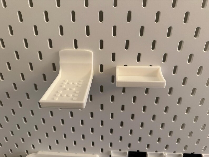 May include: Two white plastic pegboard accessories: a curved shelf with a dotted pattern and a small rectangular shelf. The items are mounted on a white pegboard with evenly spaced holes.