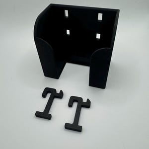 May include: Black 3D-printed wall mount with four rectangular cutouts and two black letter "T" shapes. The wall mount has a curved front and is designed to hold an item. The items are on a white surface.