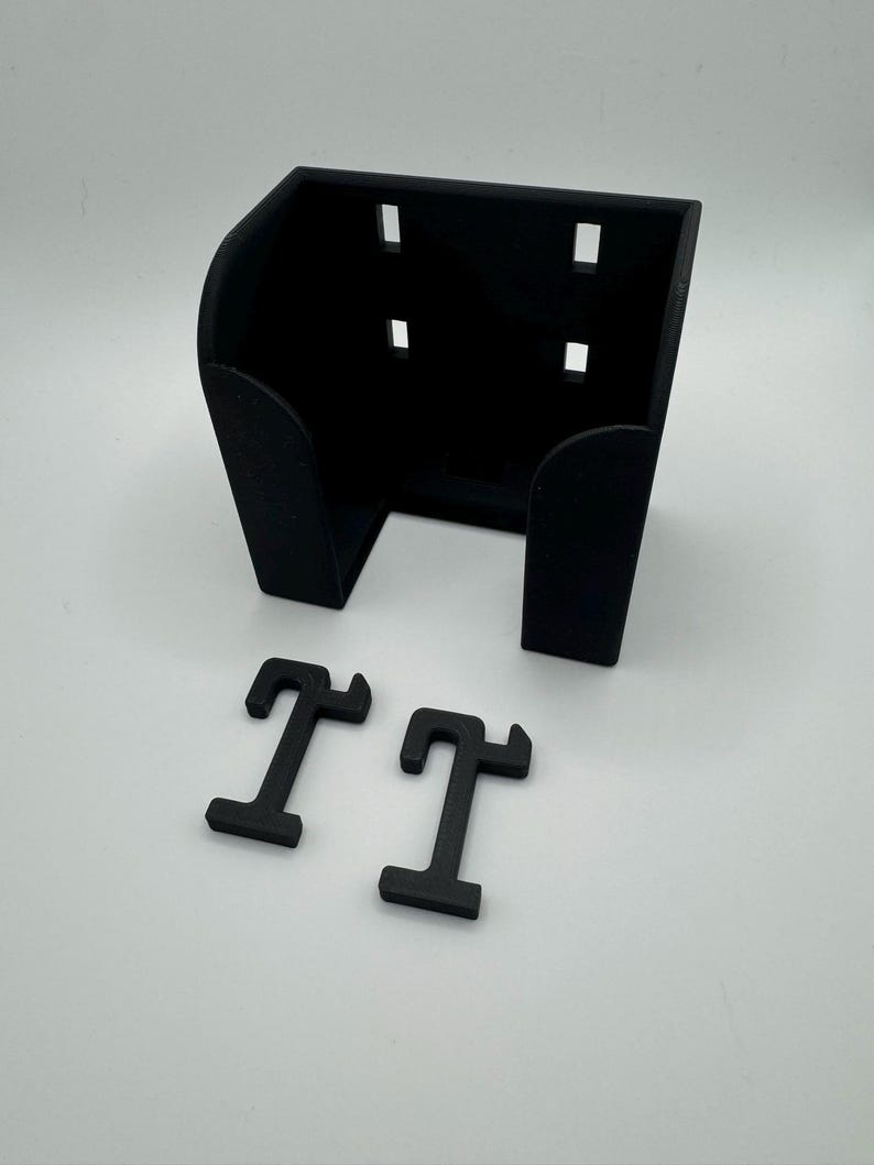 May include: Black 3D-printed wall mount with four rectangular cutouts and two black letter "T" shapes. The wall mount has a curved front and is designed to hold an item. The items are on a white surface.