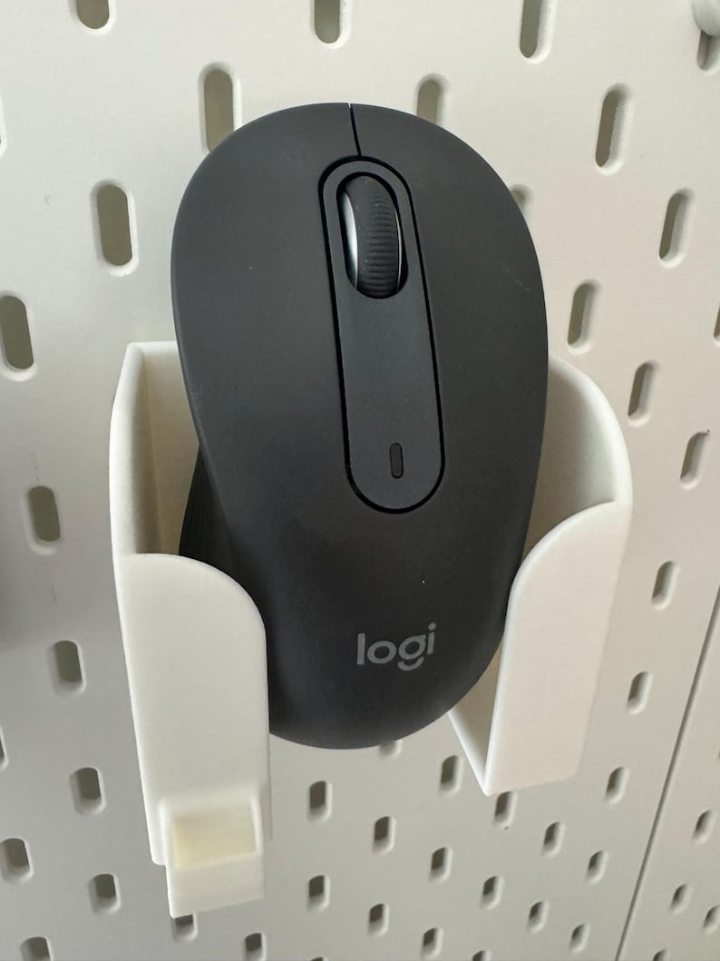 May include: A black Logitech wireless computer mouse in a white holder. The mouse has a scroll wheel and a small indicator light. The holder is mounted on a white pegboard.