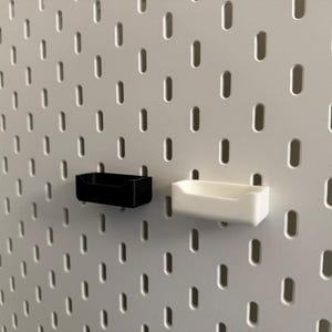 以下が含まれることがあります： 白のペグボードに、黒と白の2つの小さな長方形の収納容器が取り付けられています。容器は、小さなアイテムを収納するのに適した、シンプルなオープントップデザインです。ペグボードには、等間隔に配置された細長い穴のグリッドがあります。