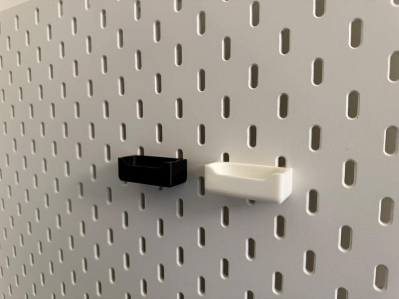 以下が含まれることがあります： 白のペグボードに、黒と白の2つの小さな長方形の収納容器が取り付けられています。容器は、小さなアイテムを収納するのに適した、シンプルなオープントップデザインです。ペグボードには、等間隔に配置された細長い穴のグリッドがあります。