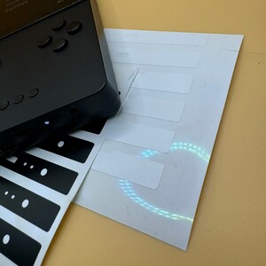 May include: A black handheld gaming device with a screen displaying the text "Play Settings", "Library", "OpenFPGA", "Memories", "Tools", and "Settings". The device has a directional pad and four buttons. The device is on a beige surface with a white sheet of paper with black stickers.