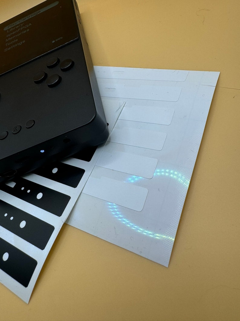 May include: A black handheld gaming device with a screen displaying the text "Play Settings", "Library", "OpenFPGA", "Memories", "Tools", and "Settings". The device has a directional pad and four buttons. The device is on a beige surface with a white sheet of paper with black stickers.